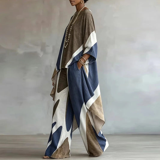 A woman models a loose, draped two-piece outfit with wide sleeves and wide-leg pants in a color-blocked abstract pattern of navy blue, taupe brown, cream, and white. She is standing in a studio with a textured gray background.