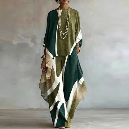 A woman models a matching two-piece outfit featuring a sage green and olive green top with white and dark green abstract color-blocked panels, paired with wide-leg trousers in a matching abstract pattern. She is accessorized with a long beaded necklace and bracelets, standing against a neutral gray studio backdrop.