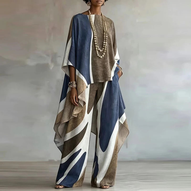 A woman models a loose, bohemian-style two-piece outfit featuring a brown textured top and matching wide-leg pants with a blue, brown, and white abstract brushstroke pattern, styled with a long pearl necklace.