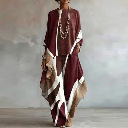 A woman models a loose, high-low two-piece outfit in a dusty burgundy, cream, and brown abstract pattern. The set includes a long-sleeve top and matching wide-leg trousers, styled with layered pearl necklaces and bracelets against a textured gray studio backdrop.