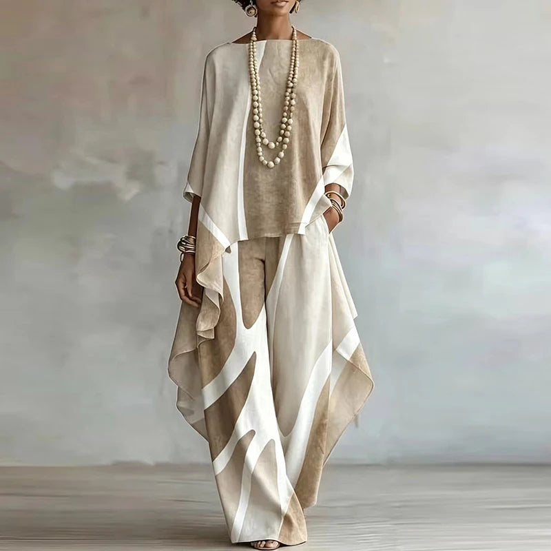 A woman models a beige and off-white two-piece linen set featuring a loose-fitting long-sleeve top and matching wide-leg trousers with an abstract draped overlay, styled with a long pearl necklace and bracelets.