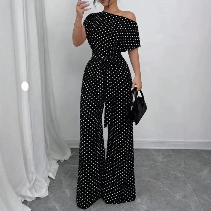 A woman models a black off-the-shoulder jumpsuit with a white polka dot pattern, featuring a tie-waist belt and wide-leg pants. She is holding a small black handbag in a room with a gray tiled floor and white curtains.