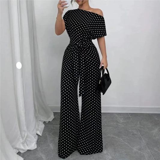 A woman models a black off-the-shoulder jumpsuit with a white polka dot pattern, featuring a tie-waist belt and wide-leg pants. She is holding a small black handbag in a room with a gray tiled floor and white curtains.