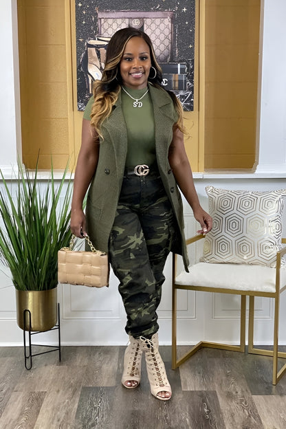 Woman in green vest and camouflage pants standing in a stylish room with decor elements.