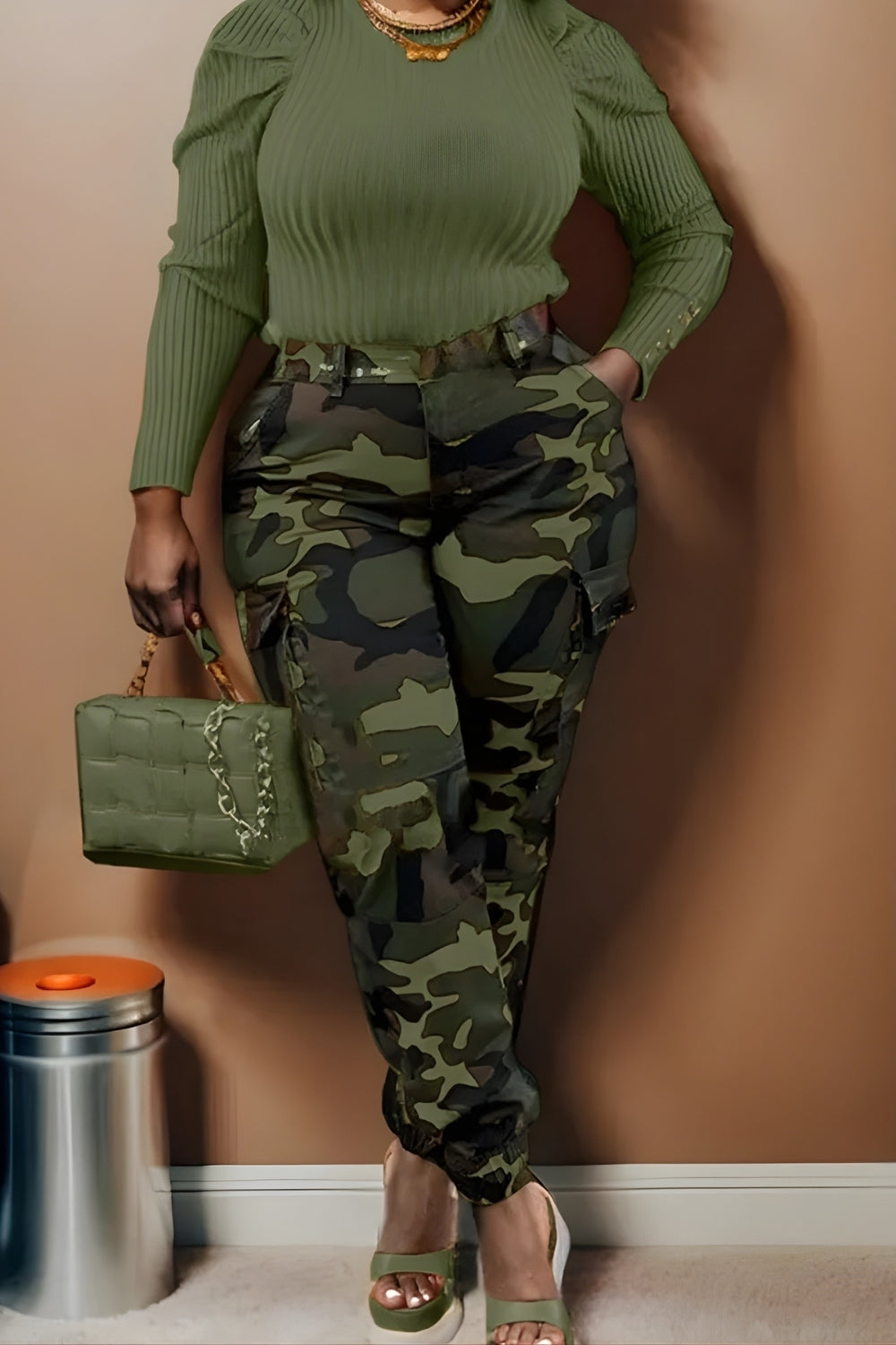 Person wearing a green ribbed top and camouflage pants, holding a green handbag against a beige wall.