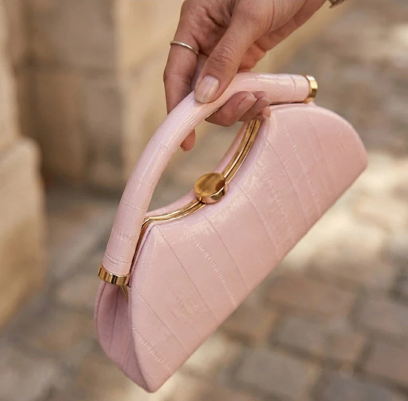 A hand holding a light pink crocodile-embossed handbag with a curved top handle and gold-tone hardware, set against a blurred outdoor stone background.