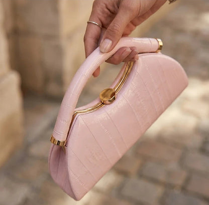 A hand holding a light pink crocodile-embossed handbag with a curved top handle and gold-tone hardware, set against a blurred outdoor stone background.