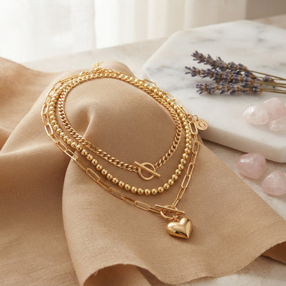 A layered gold-tone necklace set displayed on a beige fabric surface, featuring a curb chain, a beaded chain, and a paperclip chain with a small heart charm. The background includes a marble slab with lavender and pink stones.