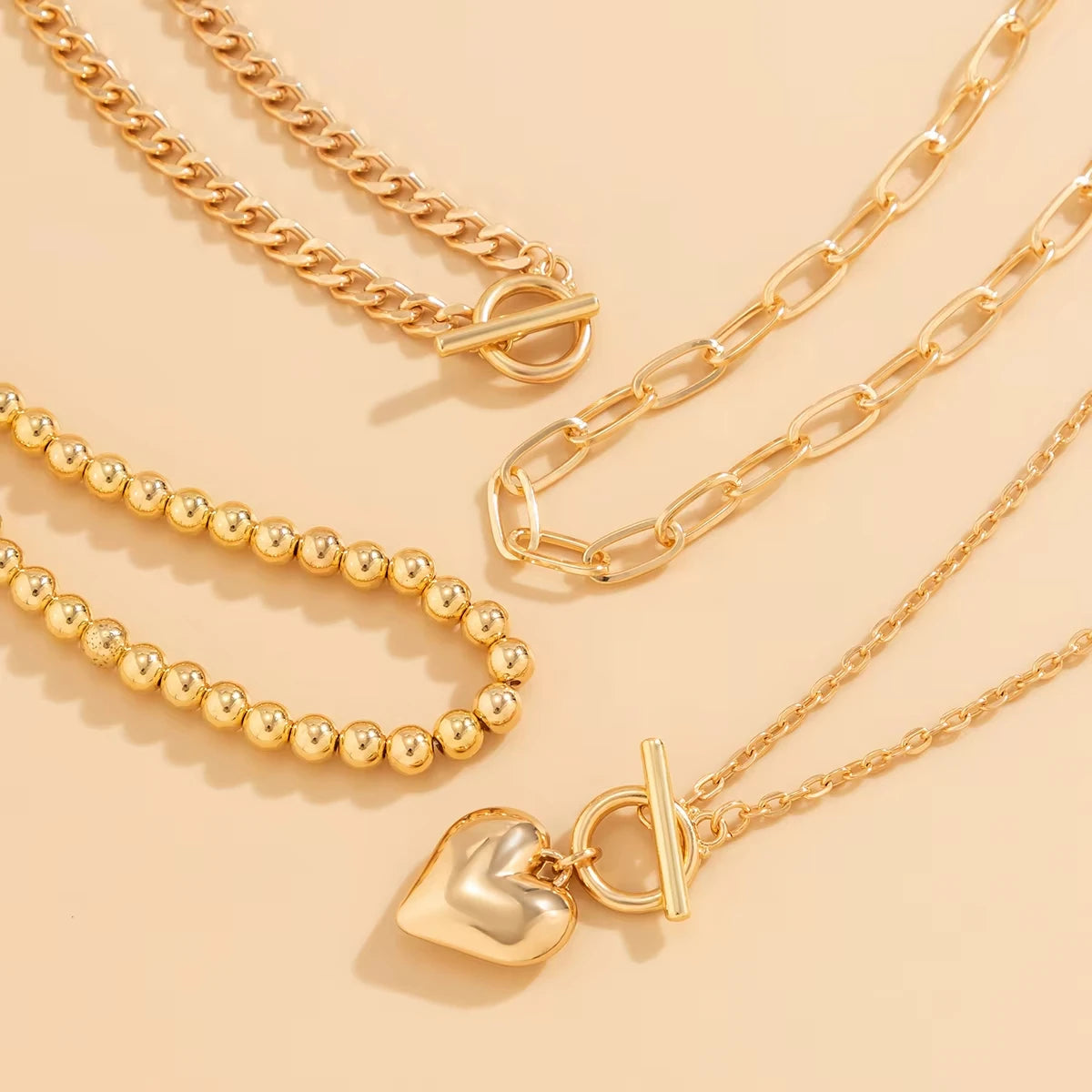 Three gold-toned jewelry pieces arranged on a warm beige background: a chunky chain necklace with a toggle clasp, a paperclip chain necklace, and a delicate chain necklace with a polished heart charm and toggle clasp.