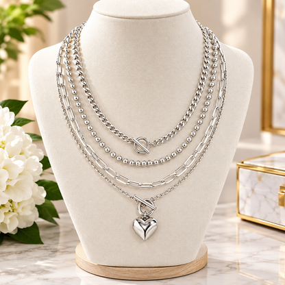A layered silver necklace displayed on a cream jewelry bust. The necklace features multiple chain styles including curb, beaded, and paperclip links, with a heart-shaped pendant hanging from a toggle clasp.