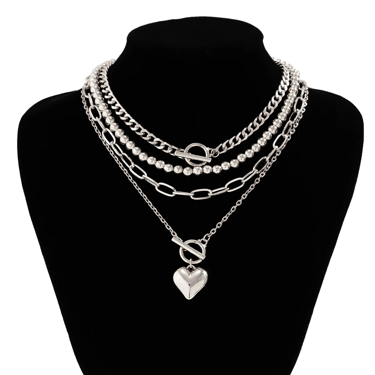 A layered silver necklace set displayed on a black neck mannequin. The set includes a chunky curb chain, a beaded chain with a toggle clasp, an oval-link chain, and a delicate chain with a heart-shaped pendant.