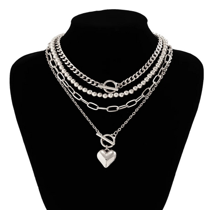 A layered silver necklace set displayed on a black neck mannequin. The set includes a chunky curb chain, a beaded chain with a toggle clasp, an oval-link chain, and a delicate chain with a heart-shaped pendant.