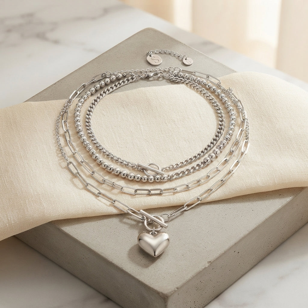A set of layered silver-tone chains with a small heart-shaped pendant, displayed on a cream fabric drape over a gray stone block.