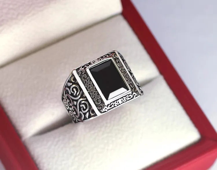 A silver signet ring with an engraved filigree band and a rectangular black stone, displayed inside a red jewelry box with a cream velvet interior.