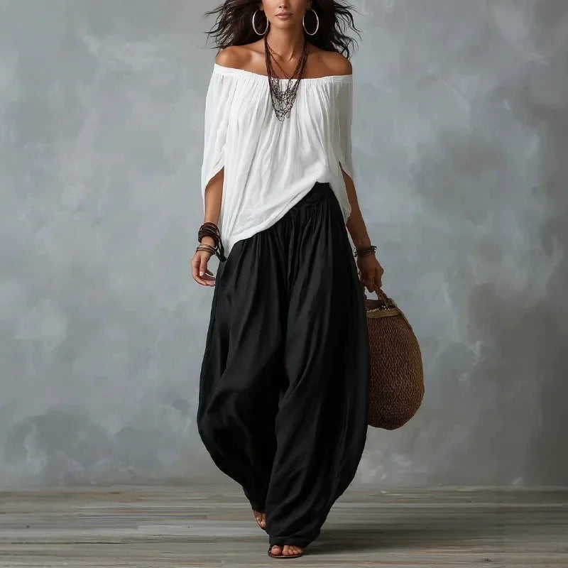 Woman wearing a white off-shoulder top and black harem pants, holding a woven bag against a gray background.