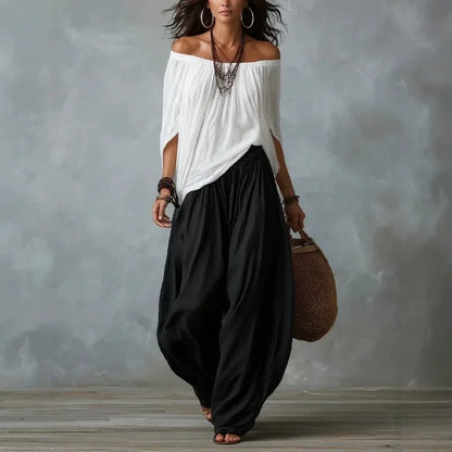 Woman wearing a white off-shoulder top and black harem pants, holding a woven bag against a gray background.
