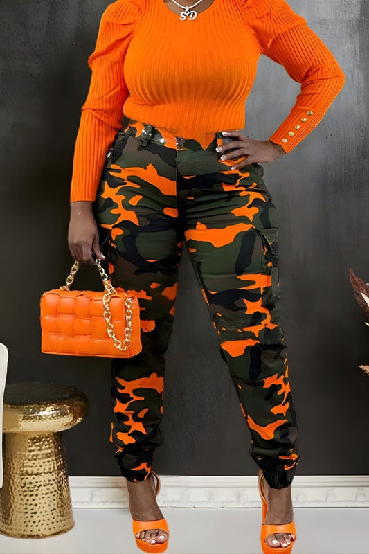 Person wearing an orange top and camouflage pants, holding an orange handbag against a black background.