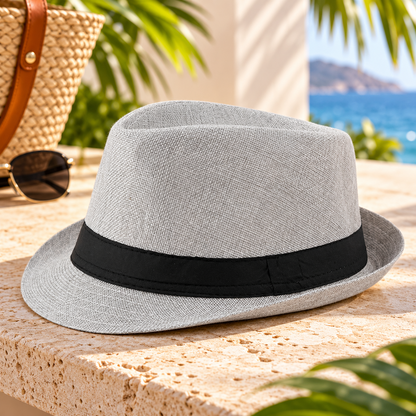 Light gray woven fedora hat with a black fabric band and short curved brim, displayed on a sunlit stone surface with sunglasses, a woven handbag, and tropical resort scenery.