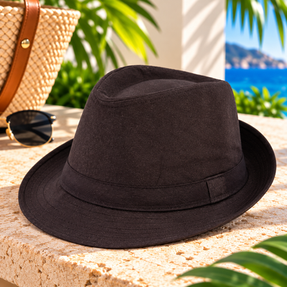 Dark espresso-brown fedora hat with a matching fabric band and short curved brim, displayed on a sunlit stone surface with a woven handbag, sunglasses, tropical greenery, and ocean scenery.