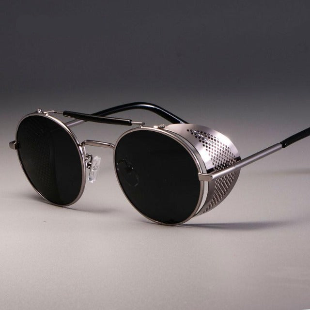 A pair of round sunglasses with silver frames, dark lenses, and perforated metal side shields, displayed on a neutral surface.